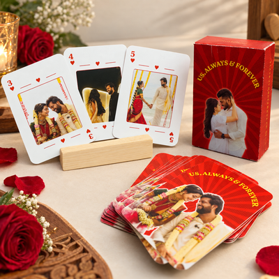 Custom wedding playing cards displayed on a tabletop, including three face-up cards showing custom couple photos, a wooden stand, a red deck box with the text 'US ALWAYS & FOREVER' and a couple photo, and a fanned stack of red cards. Red roses and floral decor surround the cards.