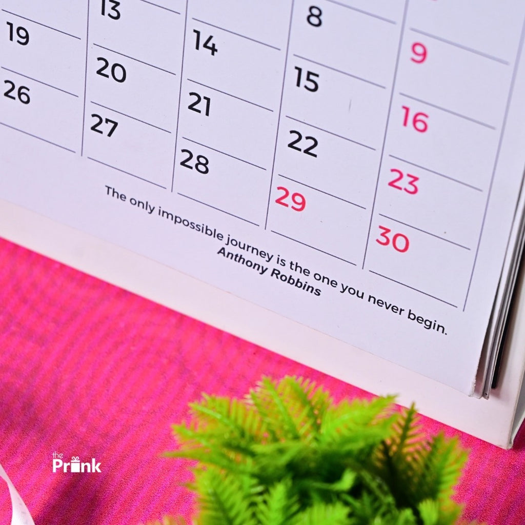 Close-up of a calendar page showing the last two weeks, including a quote by Anthony Robbins. A green plant and pink fabric are in the background.