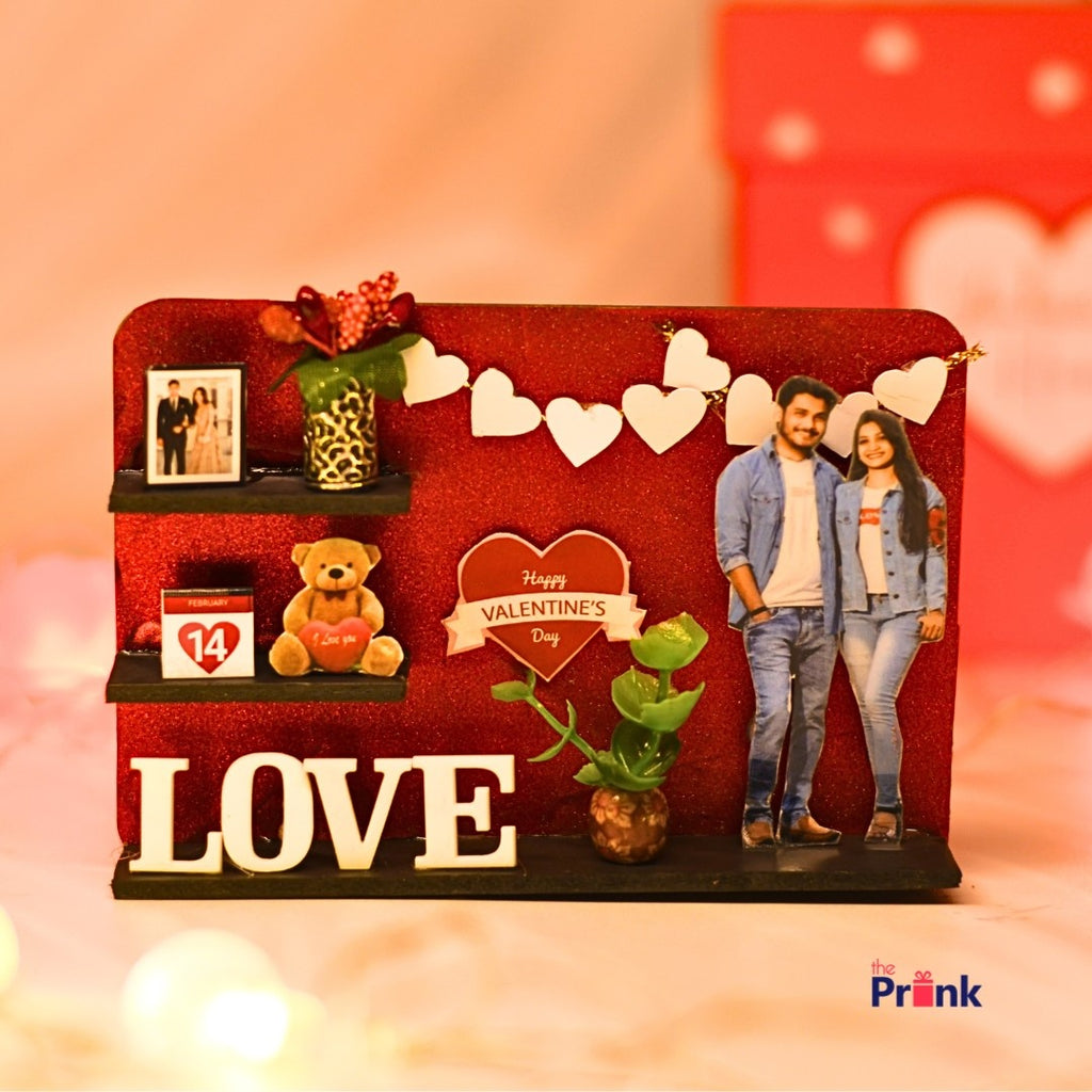 Valentine's Day-themed decor magnet with a red background features heart garland, a couple cutout, teddy bear, plants, photo, February 14th block, and "LOVE" text.