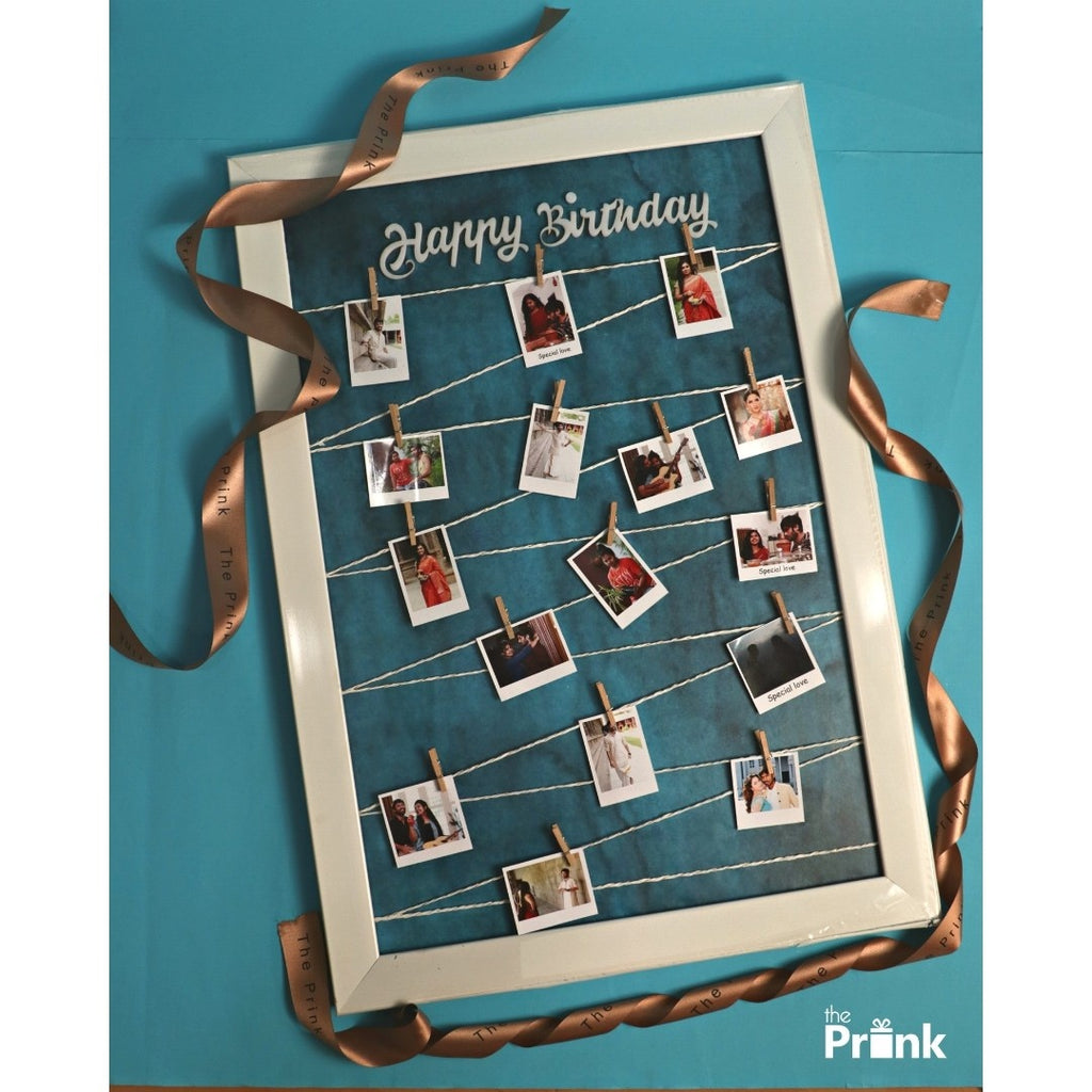 Framed photo display with the word "Happy Birthday" at the top, featuring polaroid photos clipped to white strings. Surrounding the frame are clothespins and a brown ribbon on a blue background.
