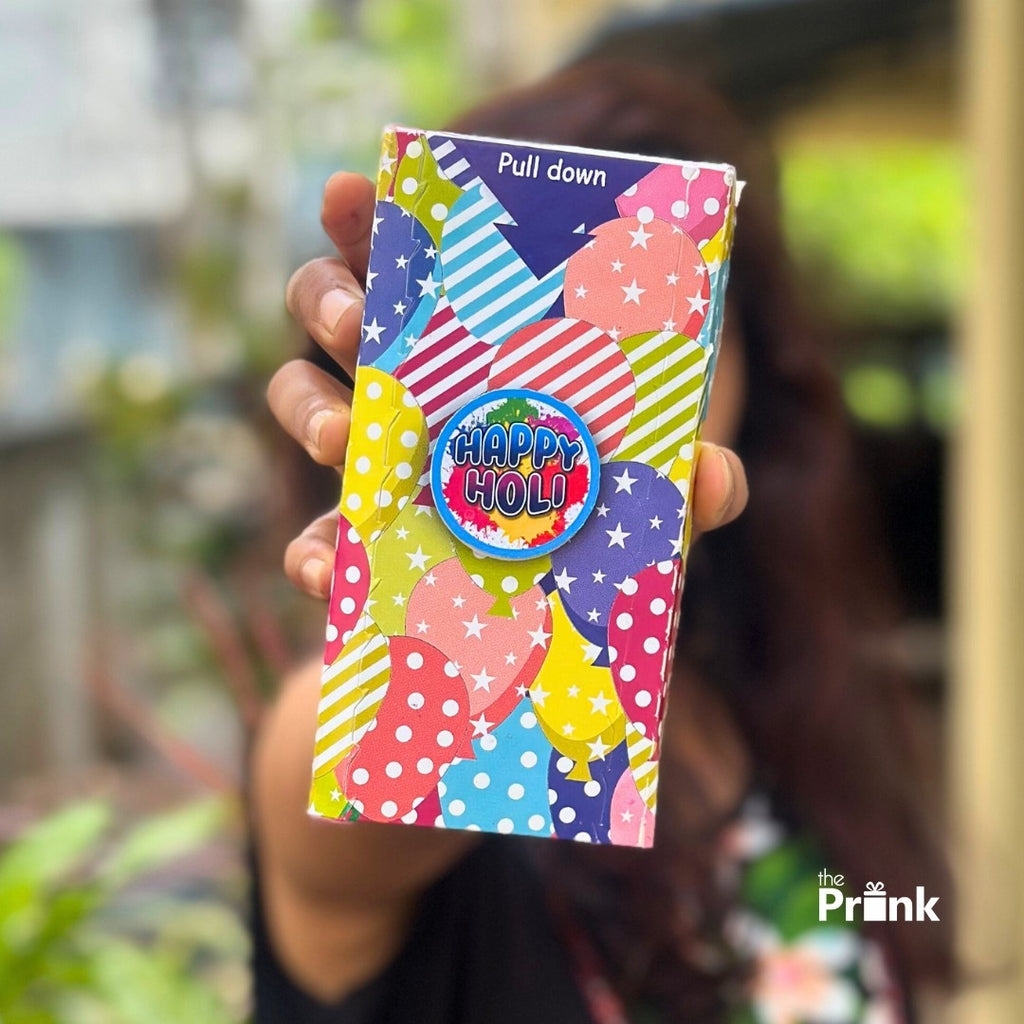 A hand holds a vibrant, colorful Holi-themed box adorned with patterns of dots and stripes, featuring a "Happy Holi" badge in the center.
