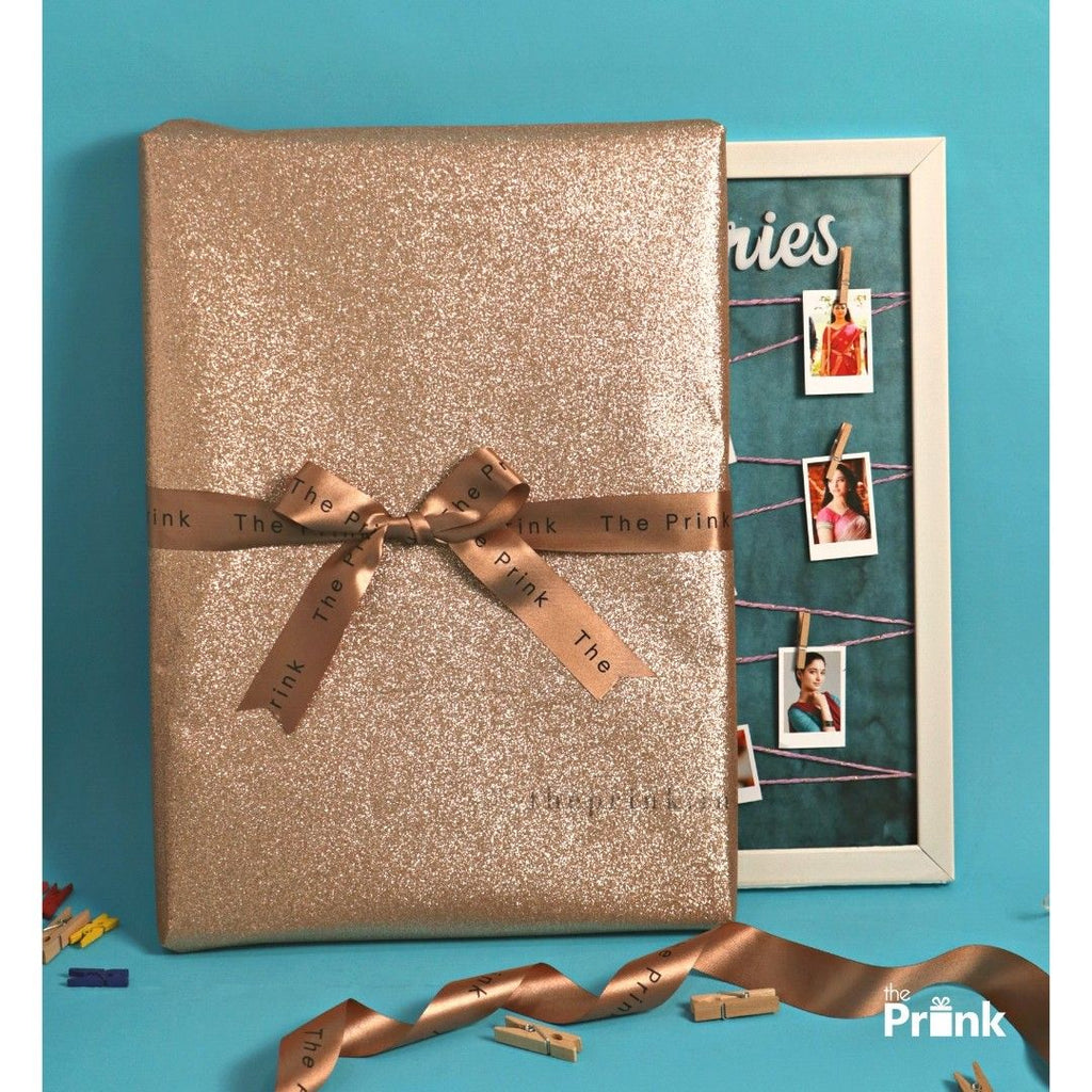 A large, glittery gold gift wrapped with a brown ribbon sits against a blue background. Behind it, a photo frame with small pictures and colorful clips adds charm.