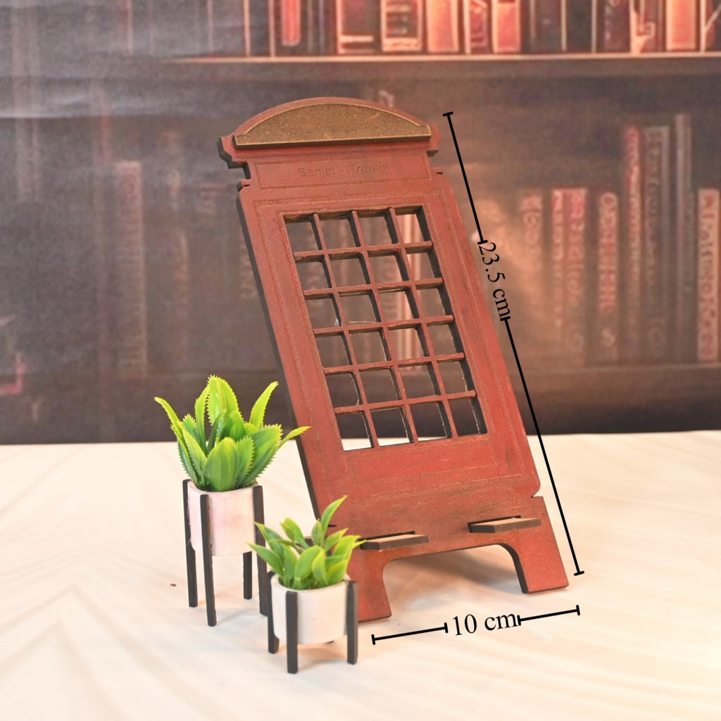 Decorative wooden phone stand resembling a vintage telephone booth, measuring 23.5 cm tall and 10 cm wide, with two small potted plants, set against a bookshelf backdrop.