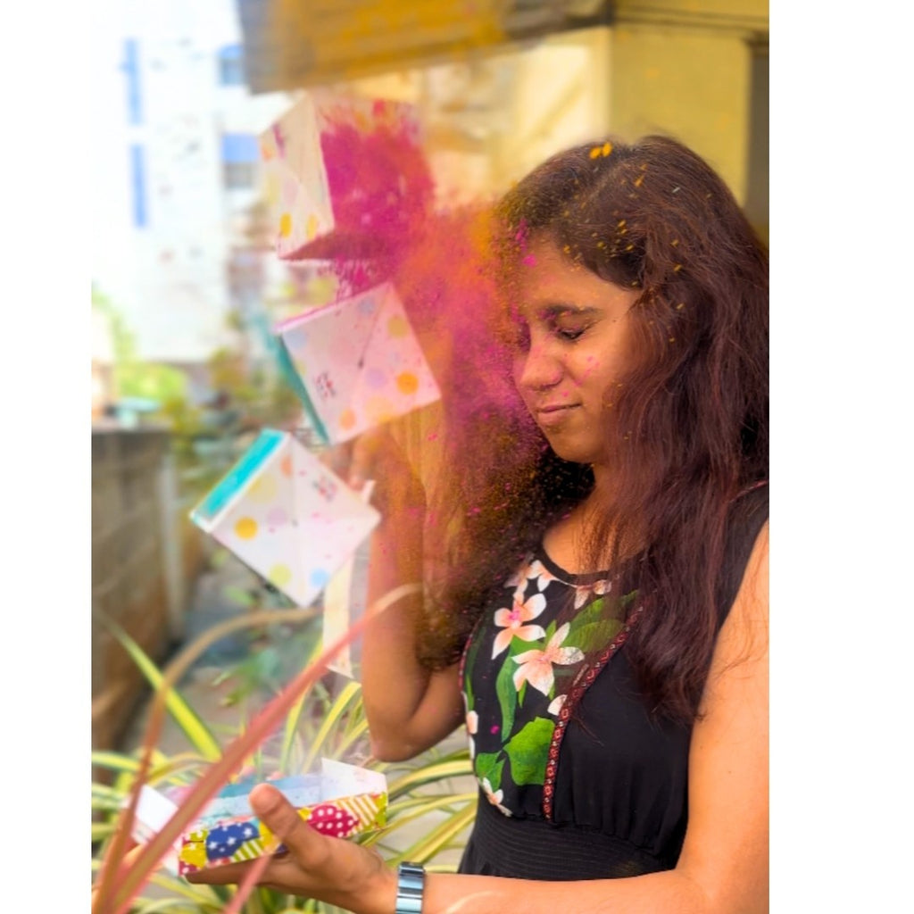 A woman on a balcony opens a colorful box, releasing vibrant pink powder and cubes into the air. Her expression is joyful.