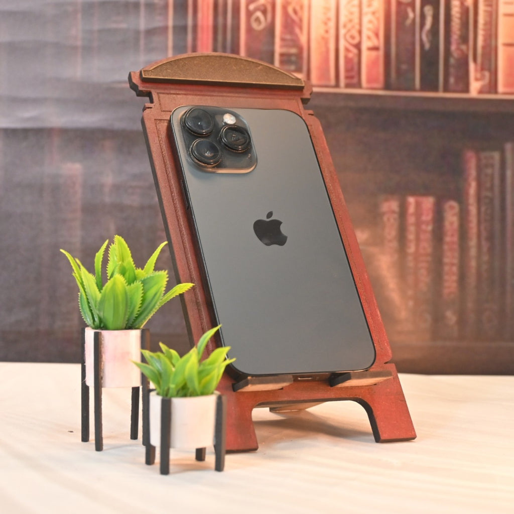 A sleek smartphone on a wooden stand, flanked by two small potted green plants. Background features blurred bookshelves, creating a cozy feel.