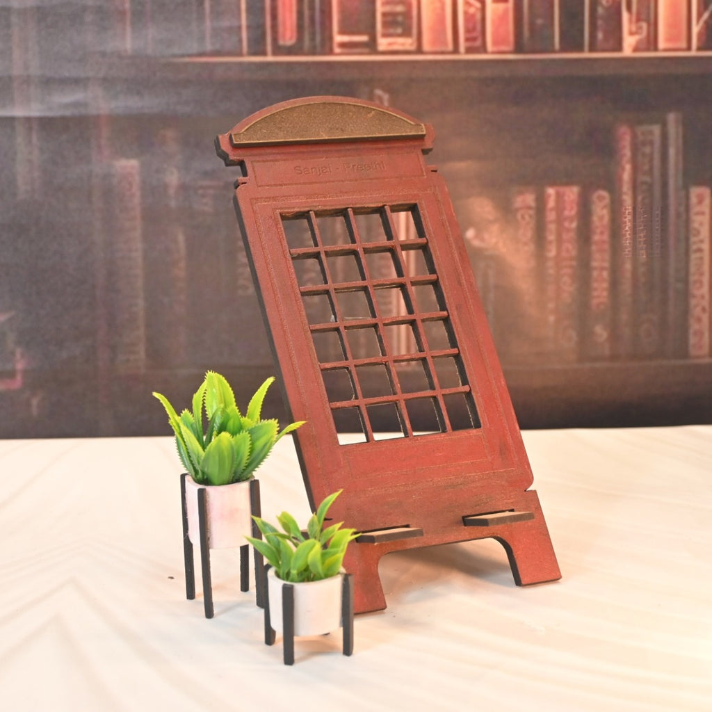 Decorative wooden phone stand resembling a vintage telephone booth, measuring 23.5 cm tall and 10 cm wide, with two small potted plants, set against a bookshelf backdrop.