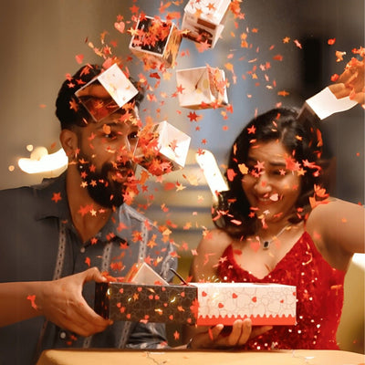 A couple dressed stylishly opens a gift box, causing star-shaped confetti to burst out joyfully. Both display excited, surprised expressions.