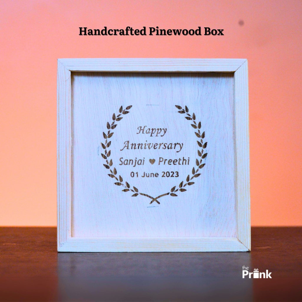 A handcrafted pinewood box with engraved text: "Happy Anniversary Sanjai ♥ Preethi, 01 June 2023," framed by decorative laurel leaves. Warm, celebratory tone.