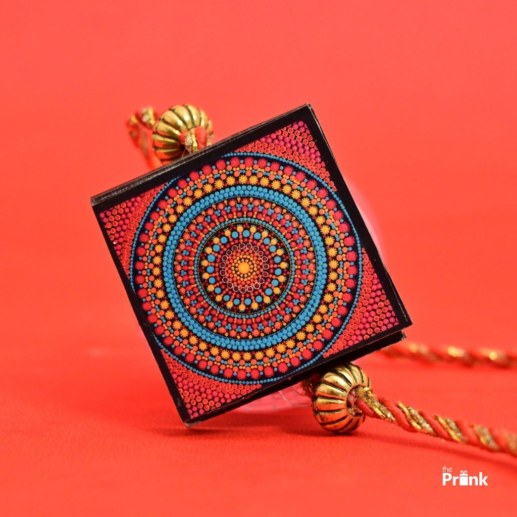 Square shaped Rakhi with personalized photos inside and intricate dot mandala design in red, blue, and yellow on a red background. Features gold accents with a vibrant, artistic tone.
