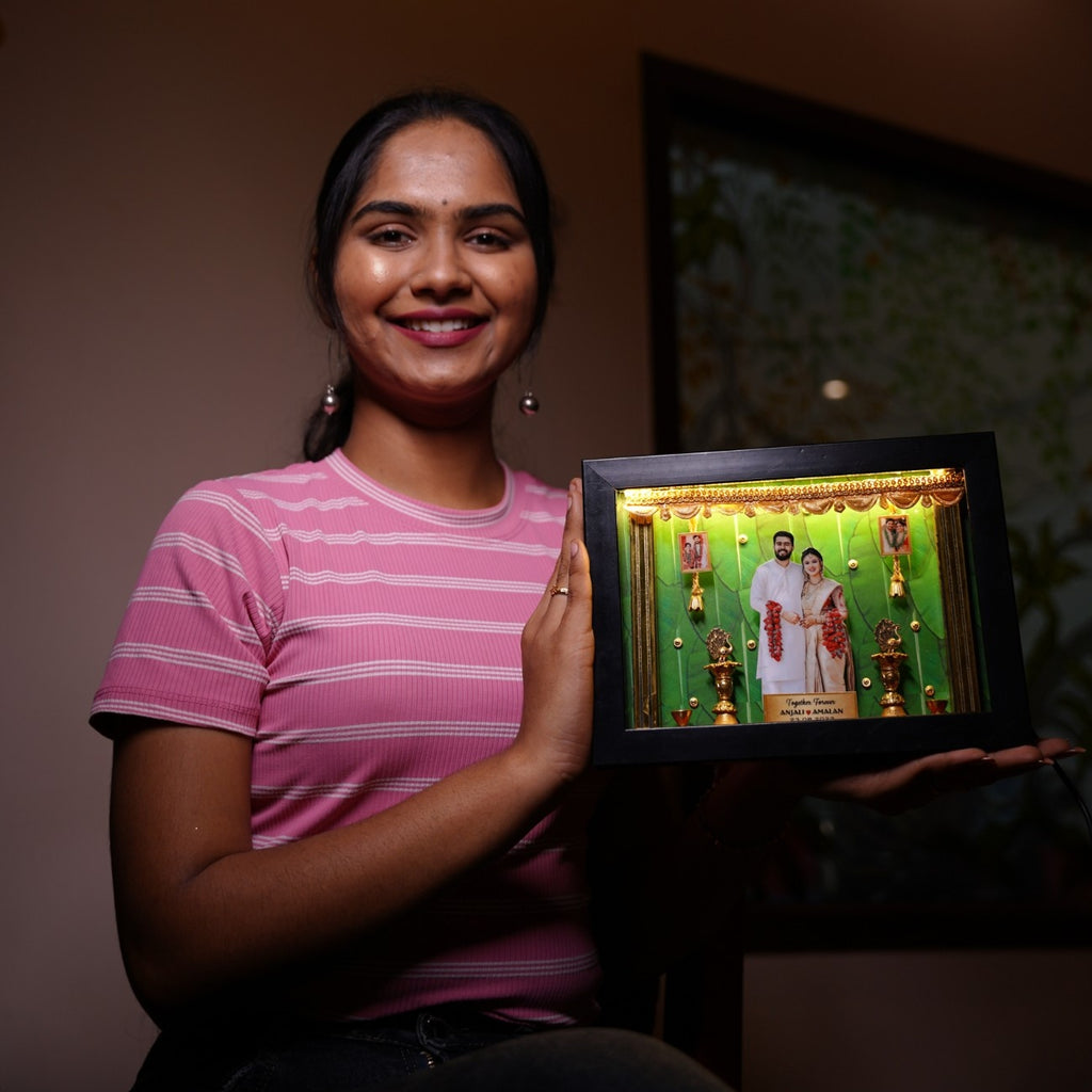 A smiling woman in a pink striped shirt holds a framed, illuminated wedding photo. The frame is ornate with gold decorations set against a green background.