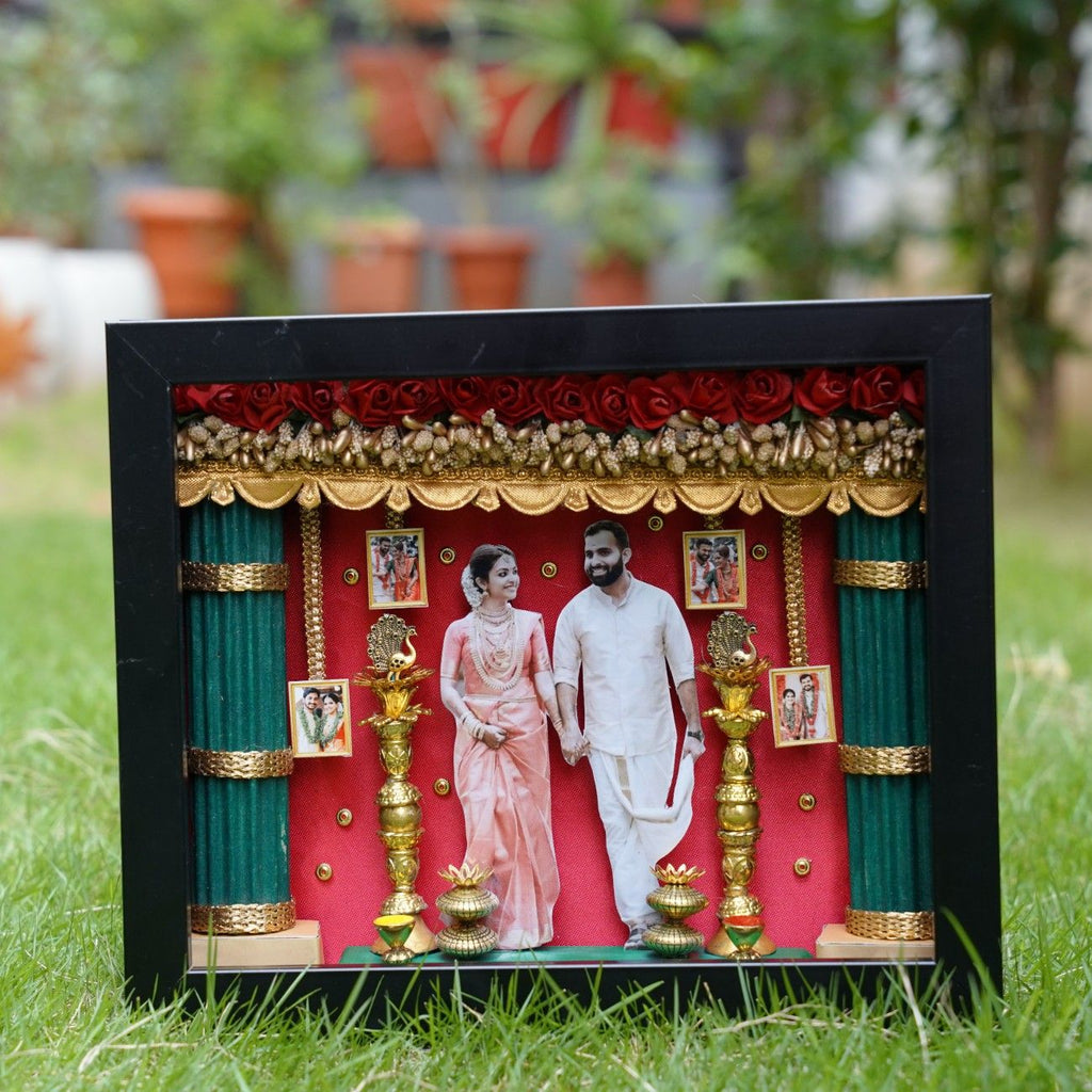 Shadow box on grass featuring a couple in traditional attire, surrounded by ornate gold decorations, red roses, and framed photos, creating a festive tone.