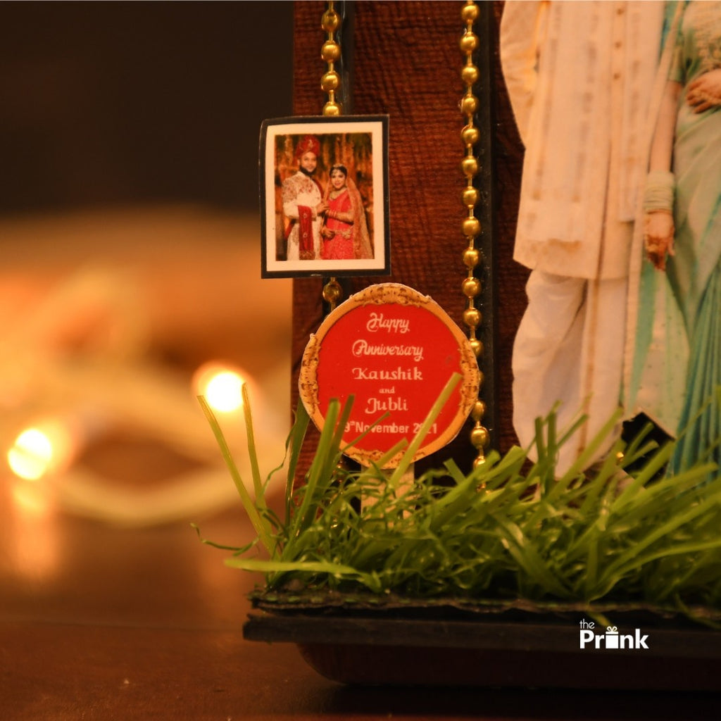 Close-up of an anniversary magnet with a small picture of a couple in traditional attire. A red circular tag reads "Happy Anniversary, Kaushik and Jubli, 3rd November 2021." Decorative beads and grass embellishments add a festive touch. Warm lighting creates a cozy, celebratory atmosphere.