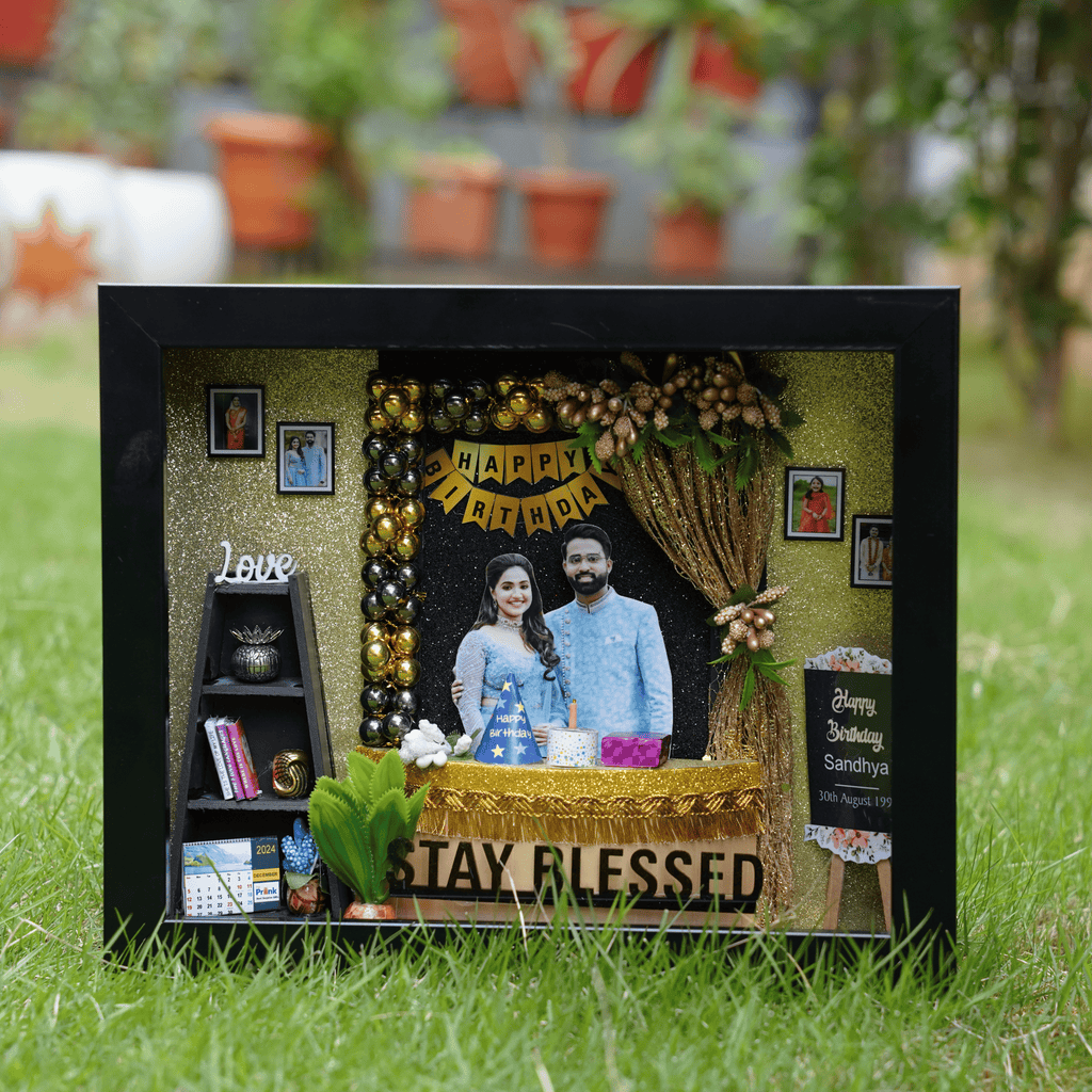 Shadow box with a couple's photo, "Happy Birthday" banner, gold accents, small shelves with books, calendar, and decorative plant, on green grass.