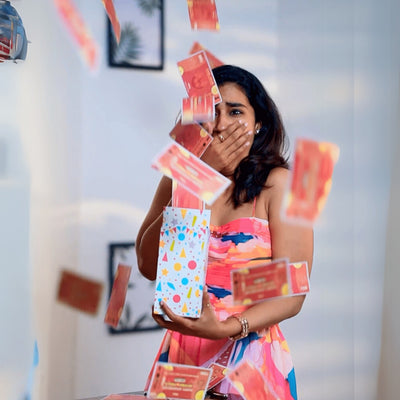 A woman in a colorful dress looks surprised as money flies out of a festive gift box. The room is bright and decorated with framed pictures.