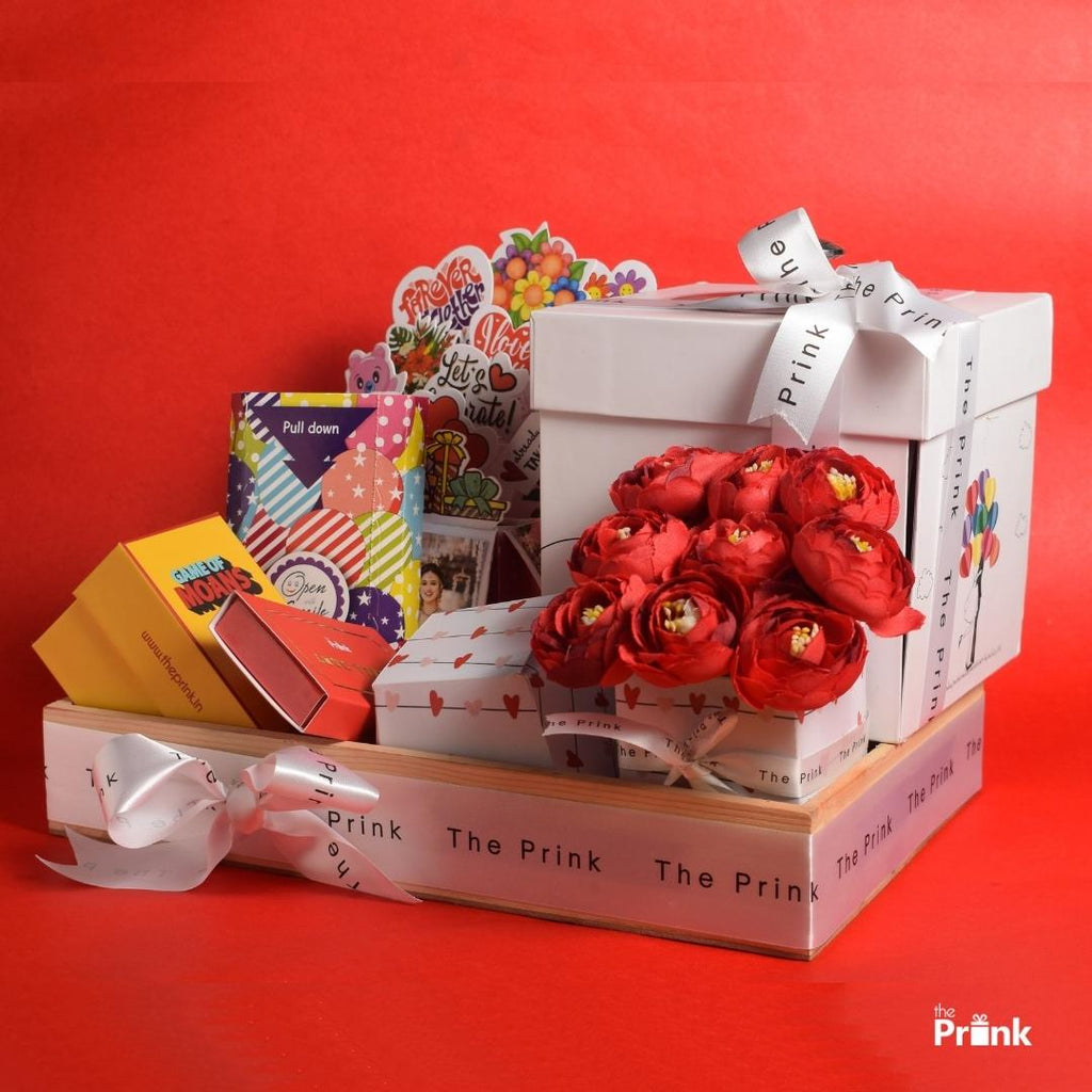 A festive gift arrangement on a red background features a large white box with a ribbon, red paper flowers, colorful cards, and smaller boxes, conveying celebration.