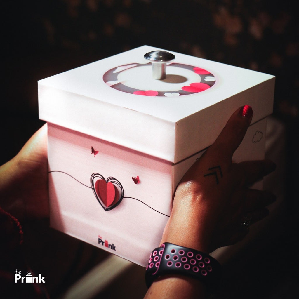 Two hands hold a white and pink gift box with heart designs and a silver knob on top. The scene has a warm, intimate, and festive feel.
