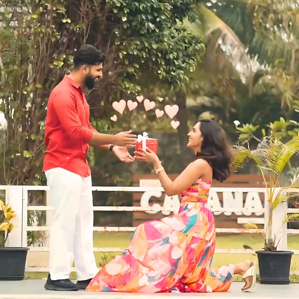 A woman in a colorful dress kneels, offering a gift box with hearts floating around. A man in a red shirt and white pants smiles, reaching for it. Romantic garden setting.