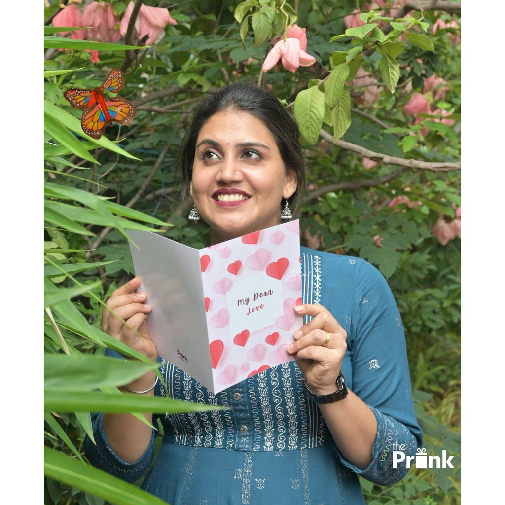 A person in a blue dress holding a card reading "My Dear Love" stands amid lush greenery and pink flowers, smiling warmly at the butterfly.