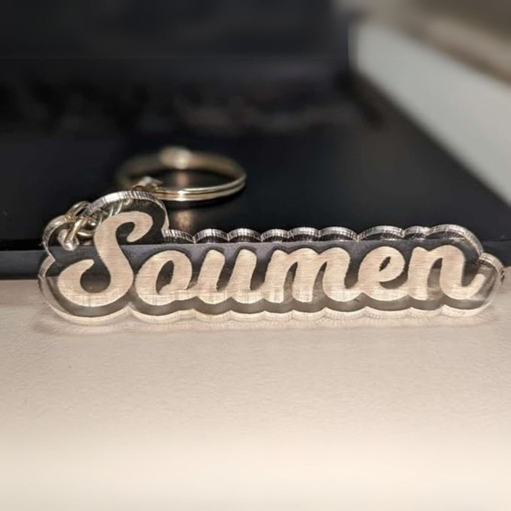 Keychain with the name "Soumen" in 3D letters, resting on a surface. The silver letters have a wavy border, conveying a sleek, personalized feel.