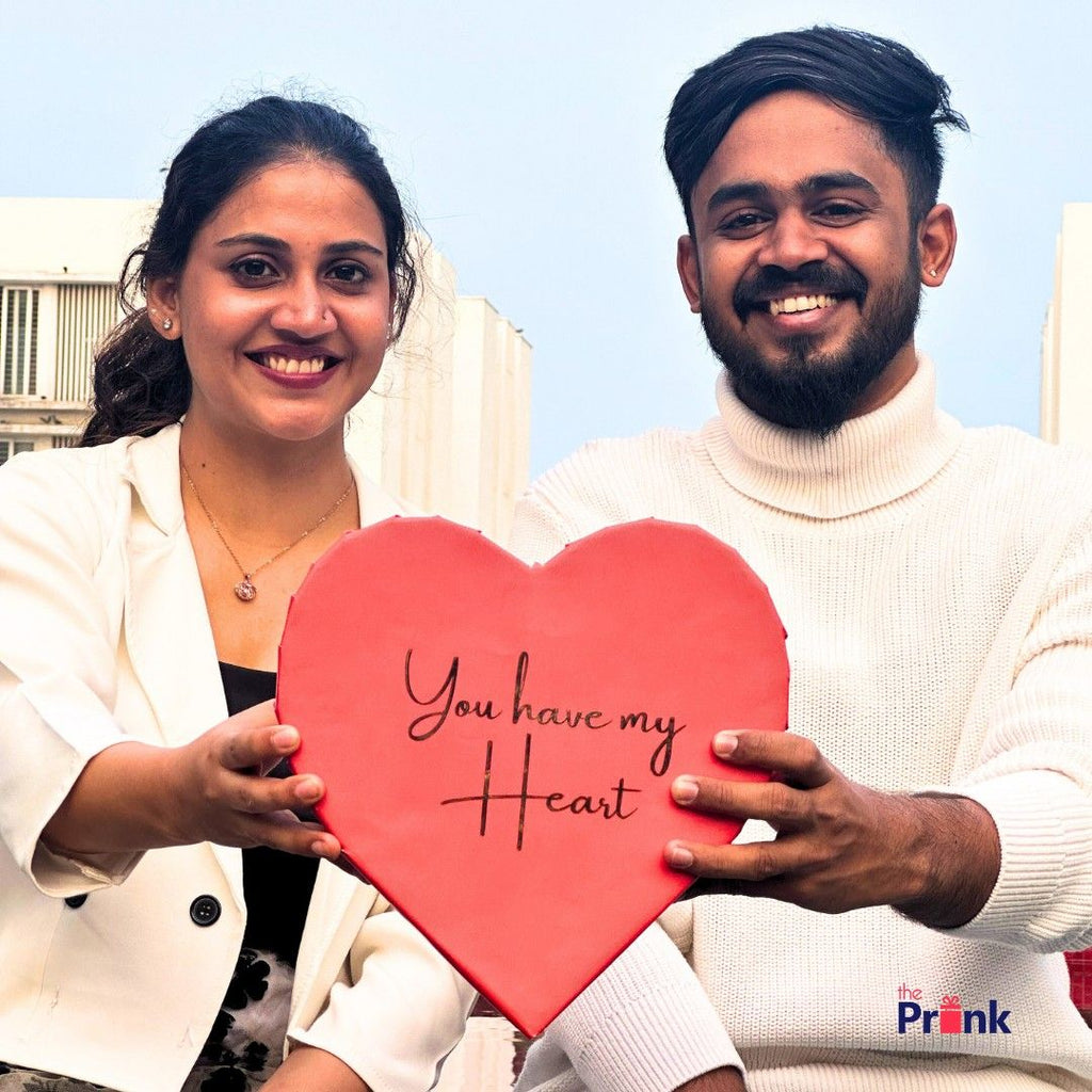 A smiling couple holds a large red heart box with "You have my Heart" written on it. They stand against a sky-blue backdrop, exuding warmth and joy.