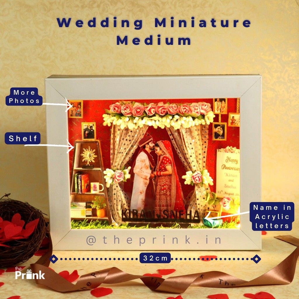 Wedding-themed shadow box with a couple's photo framed by floral garlands. Features miniature shelves with books and photos. Text includes "Kiran Sneha" in acrylic.