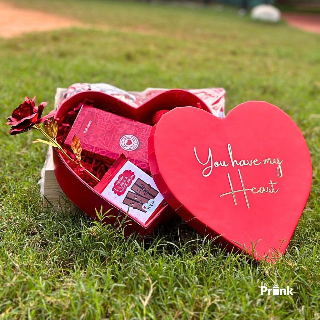 Heart-shaped red gift box on grass, lid reads "You have my Heart." Inside are chocolates, a red rose, and a romantic card, conveying love.