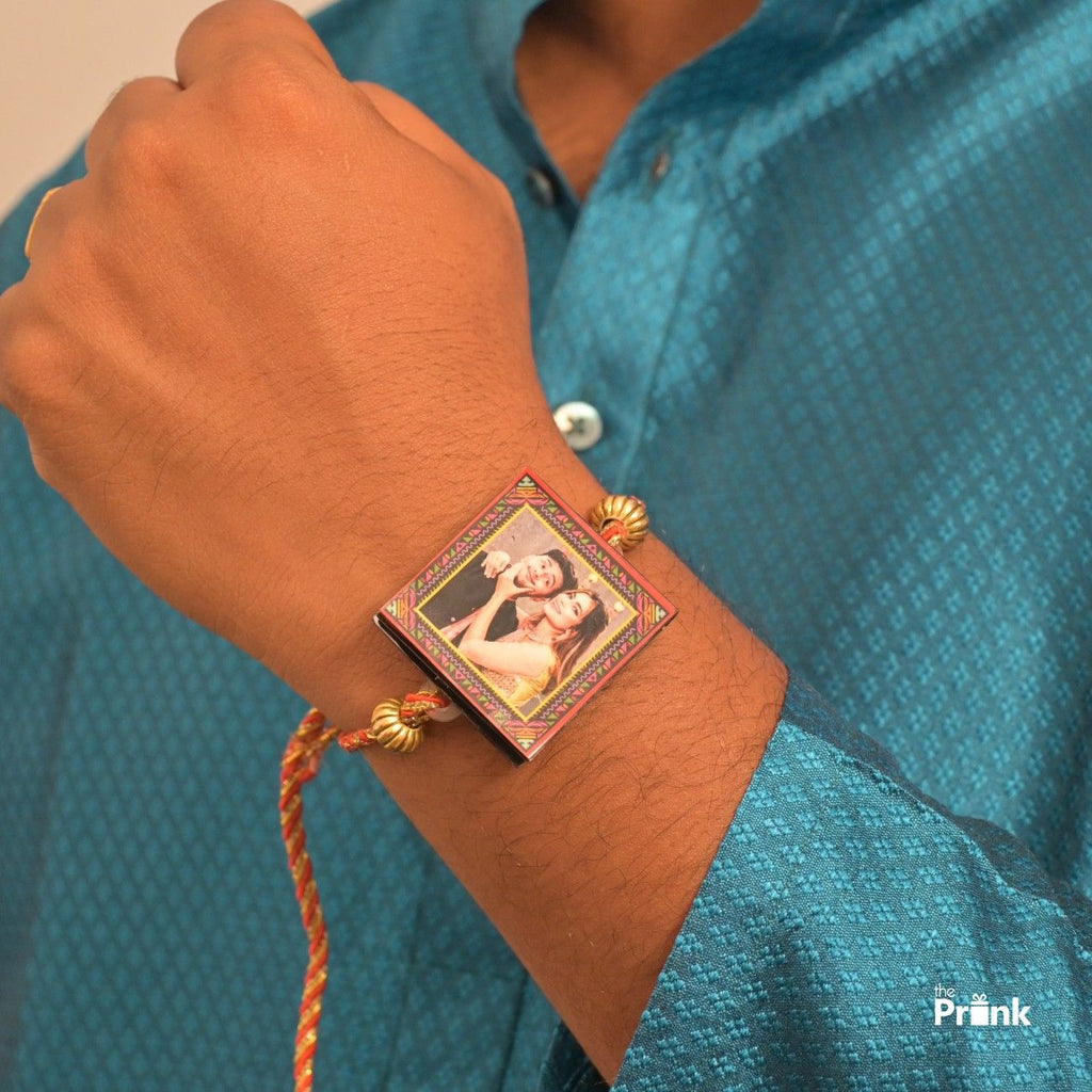 A hand wearing a rakhi bracelet with a small, framed photo of them. The bracelet has red and gold threads. The person is dressed in a blue textured shirt.