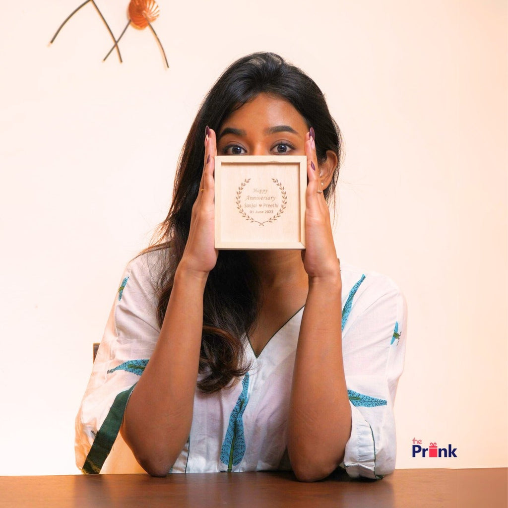 A woman in a white shirt holds a wooden plaque with engraved text, partially covering her face. The background is simple, with minimal decor.