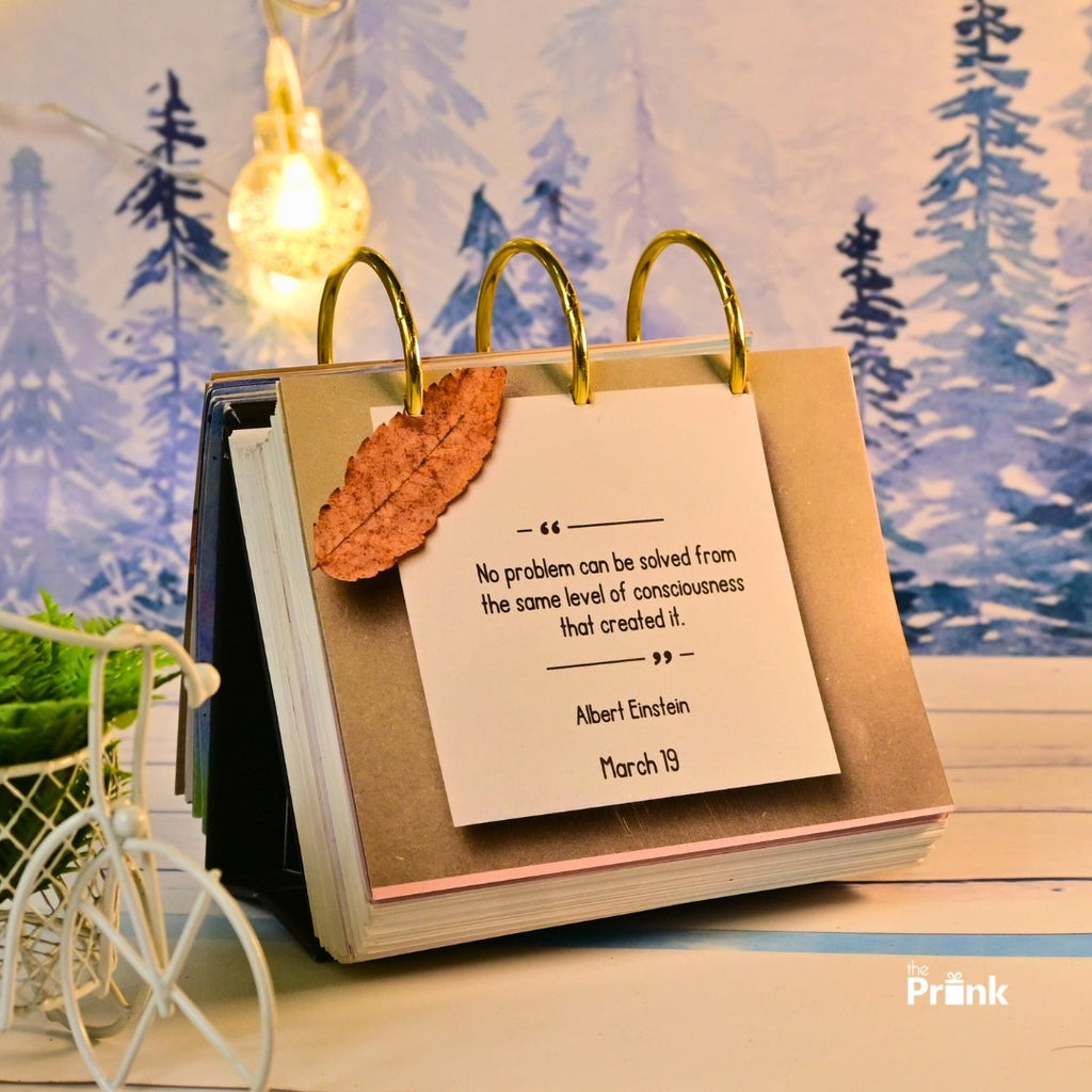 A desktop calendar displays an Albert Einstein quote on March 19, adorned with a dried leaf. Background features blue snowy trees and a glowing light bulb.