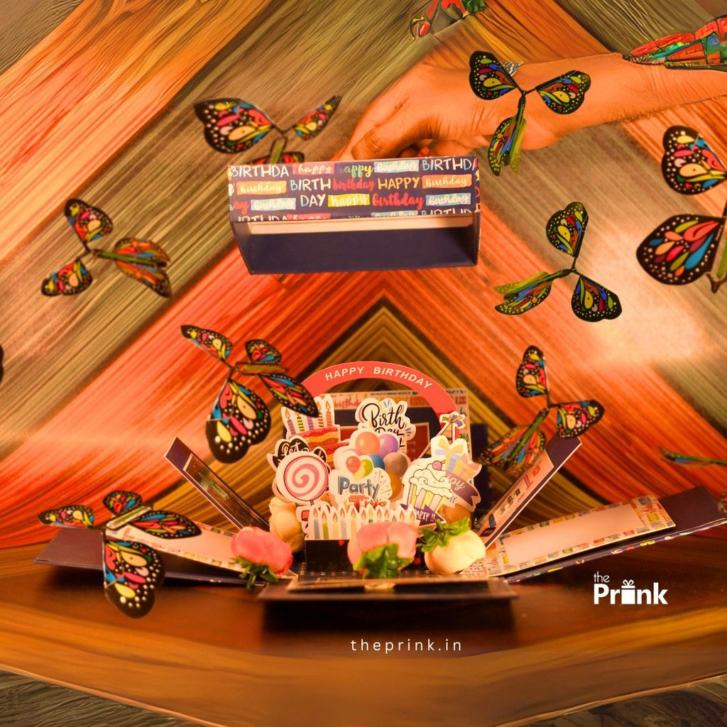 A colorful pop-up box with festive butterflies and handwritten birthday messages inside. A hand lifts the lid, revealing chocolates and roses.