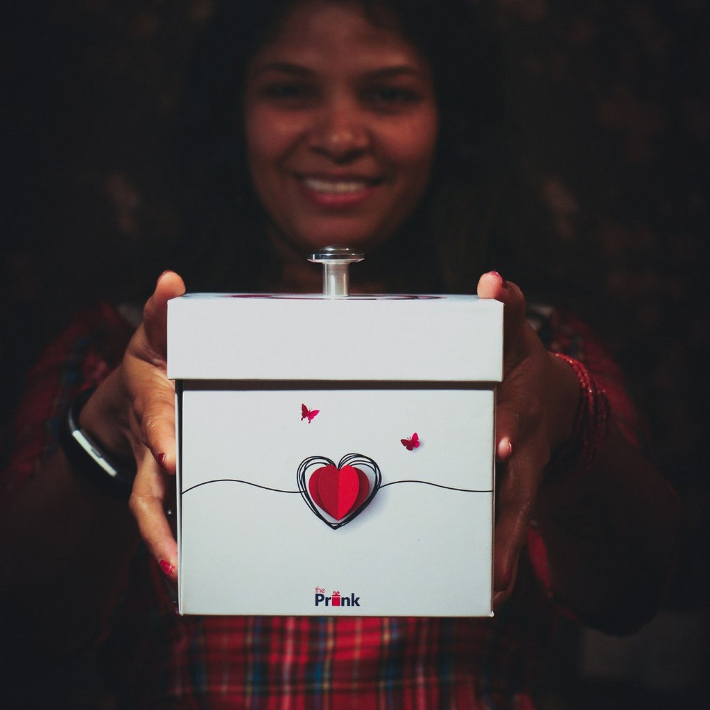 A person smiling holds a white gift box with a red heart and butterflies design. The background is blurred, creating a warm, joyful atmosphere.