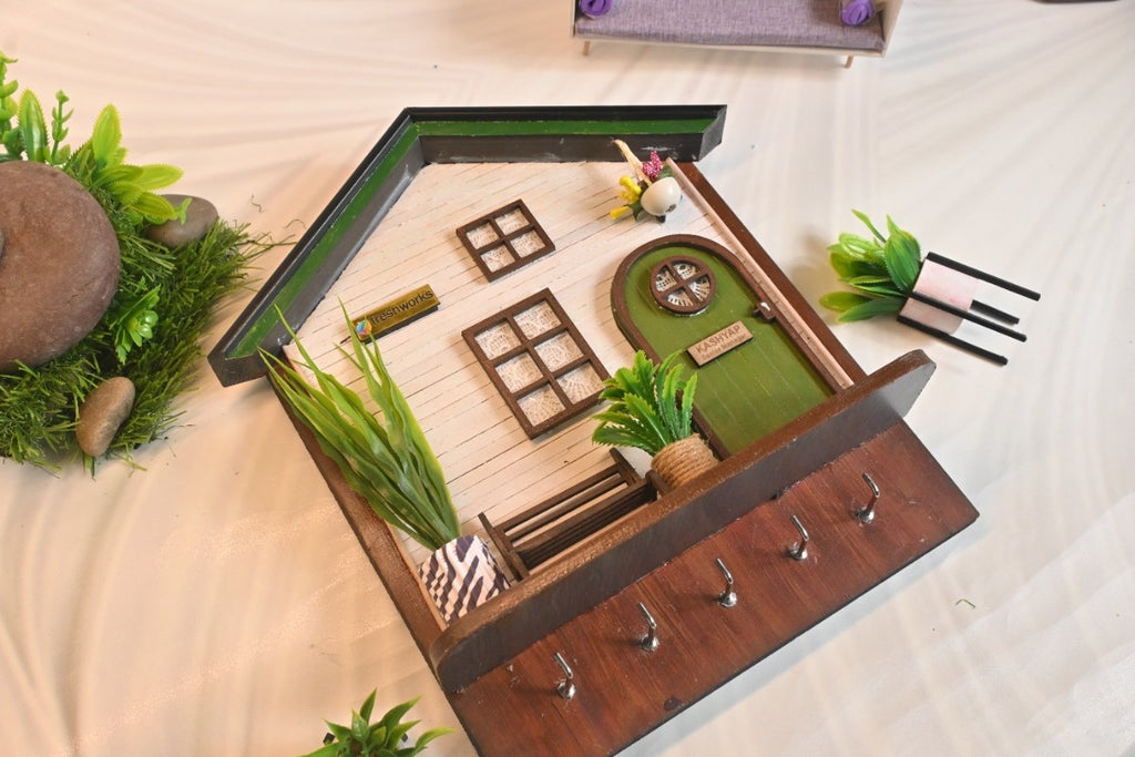 Miniature house key holder with a green door, white facade, and small windows. Decorated with faux plants and labeled "Kashyap." Five hooks are below.