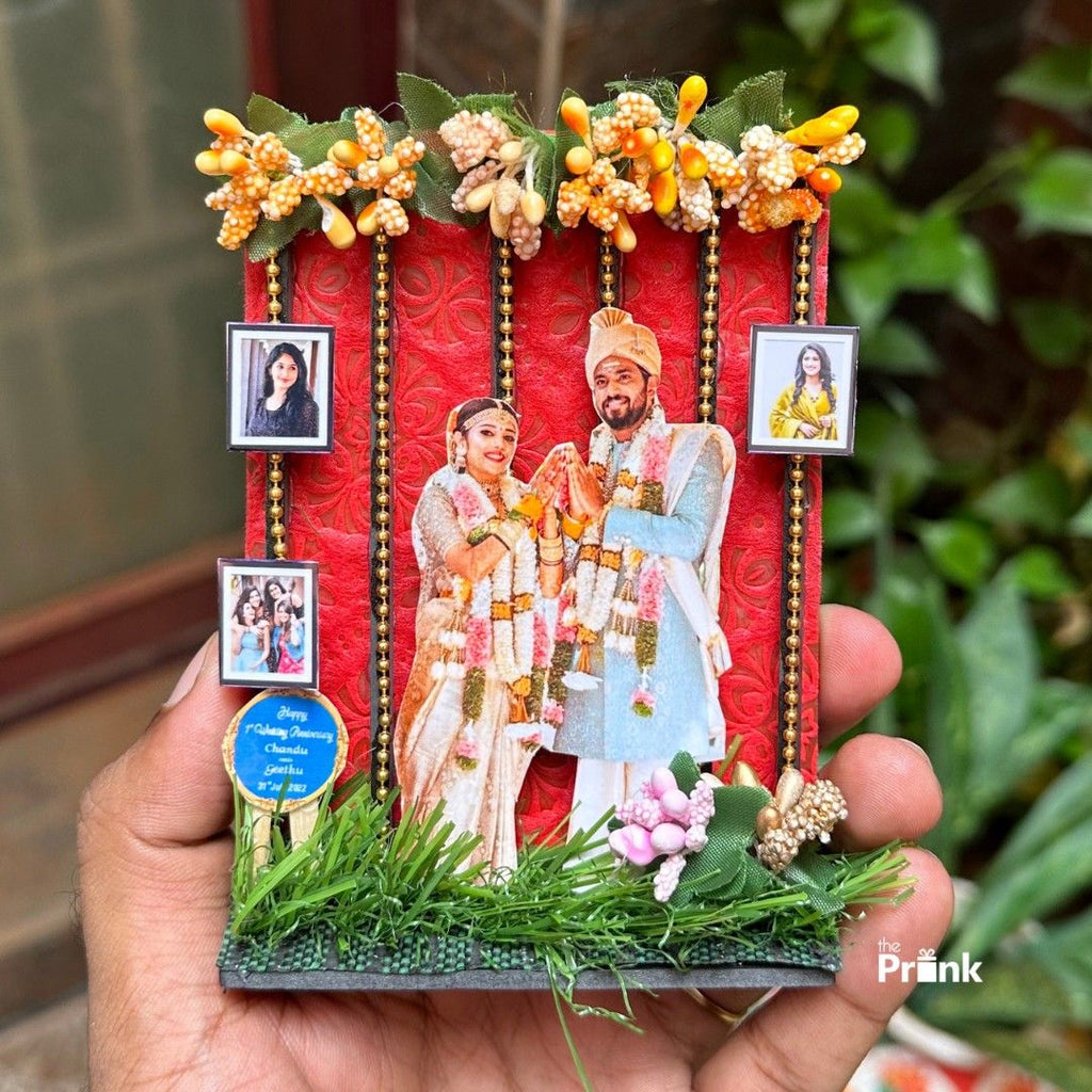 A decorative piece is held in a hand, featuring a 3D cutout of a smiling couple in traditional Indian wedding attire with floral garlands. The background is red with gold chains and topped with artificial yellow flowers. Small portrait photos and a blue circular label are visible. A green artificial grass section is at the bottom, adding to the festive and celebratory tone.
