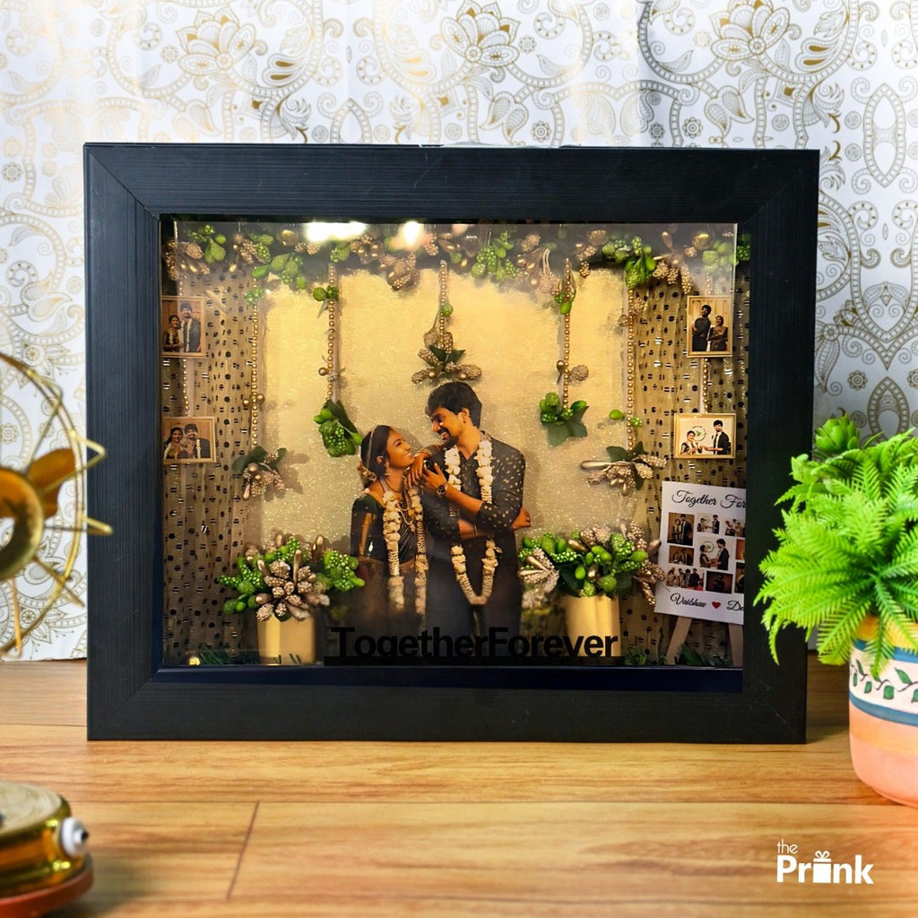 Framed shadow box with a couple dressed in festive attire, smiling fondly. Decorated with garlands, small plants, white fence, and photos, conveying joy.