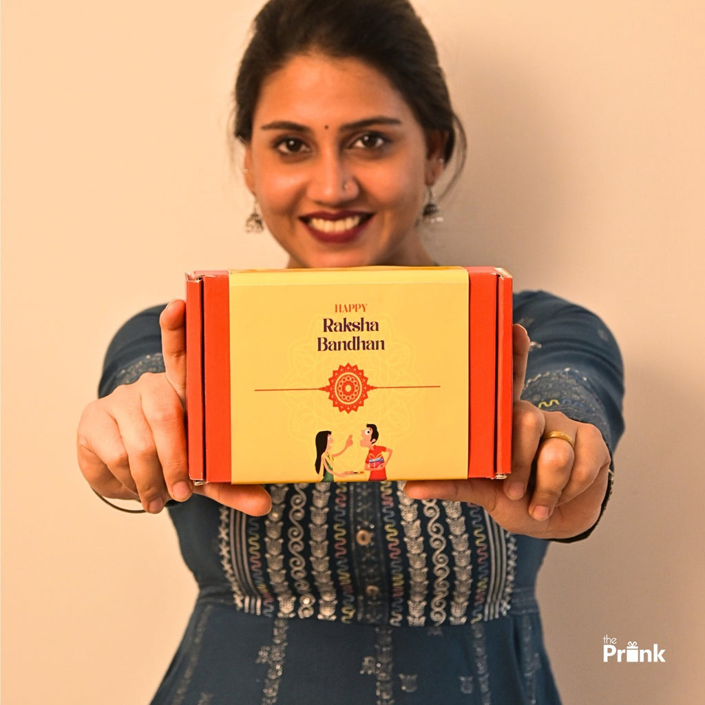 Woman in a blue dress holds a red and yellow "Happy Raksha Bandhan" gift box, smiling warmly, conveying a joyful and festive tone.