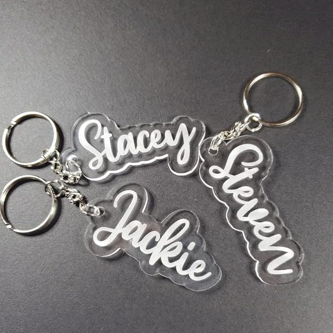 Three acrylic name keychains on a dark surface. Each keychain is shaped with names in white cursive: Stacey, Jackie, and Steven. The mood is personalized and creative.