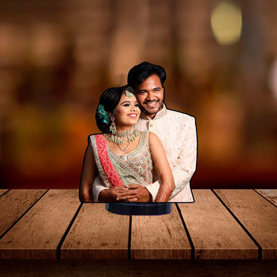 Acrylic standee featuring couple in elegant traditional attire on a surface, smiling warmly amidst a soft-focus, warmly lit background. Romantic and joyful tone.