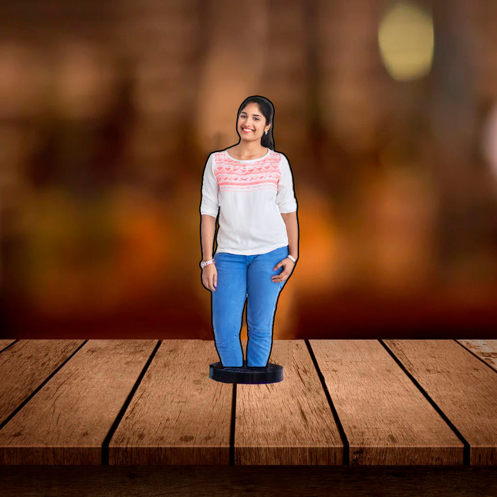 An acrylic standee of smiling woman in a white top and blue jeans placed on a wooden surface against a blurred warm-toned background.