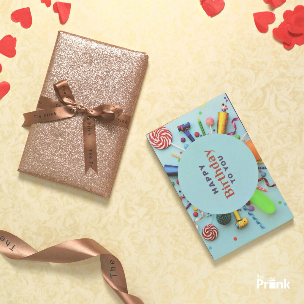A glittery, bronze gift wrapped with a satin "The Prink" ribbon, alongside a colorful "Happy Birthday To You" photo album on a light floral background. Red heart confetti scattered around, exuding a festive vibe.