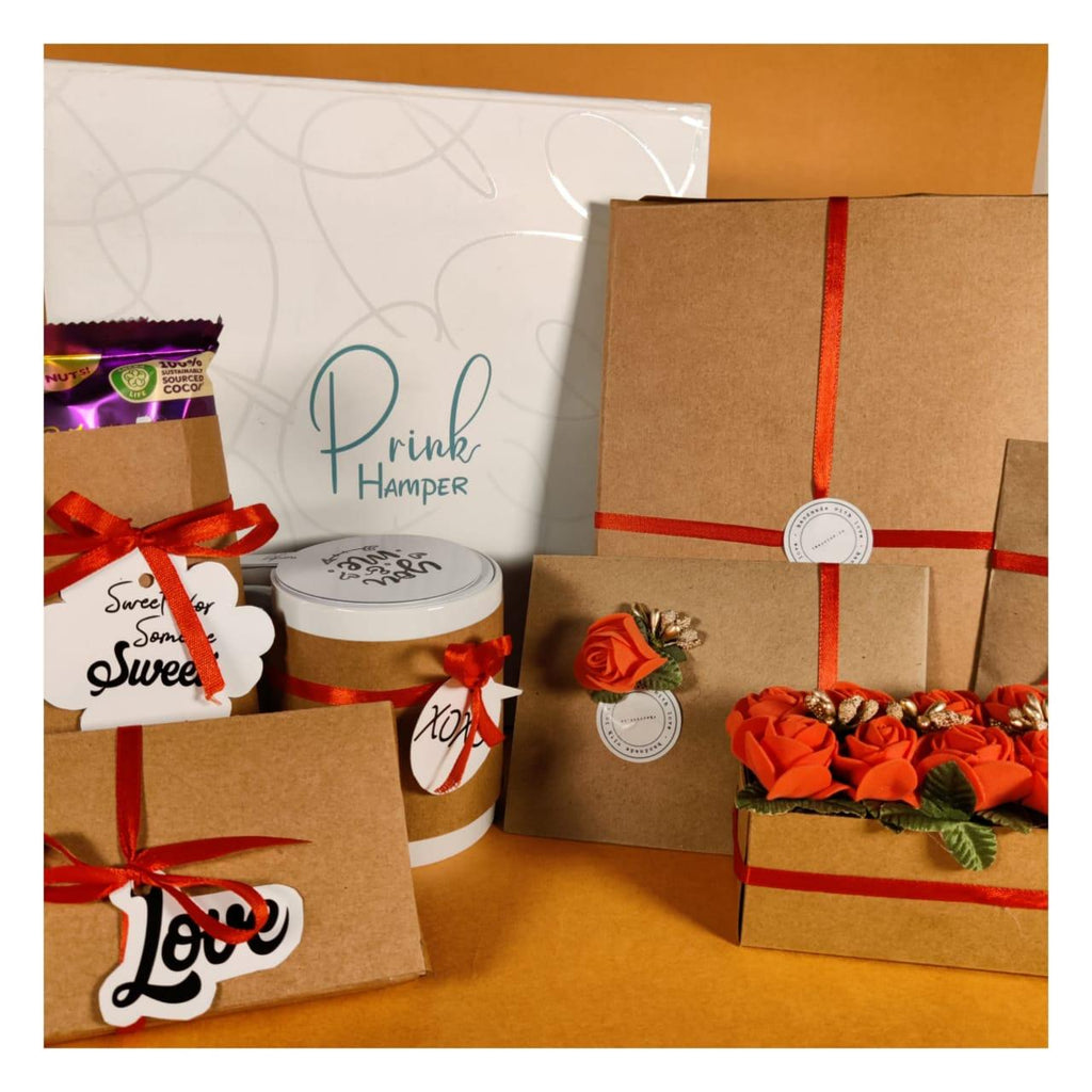 Brown gift boxes adorned with red ribbons and orange flowers are arranged on an orange surface. The background features a sign reading "Prink Hamper."
