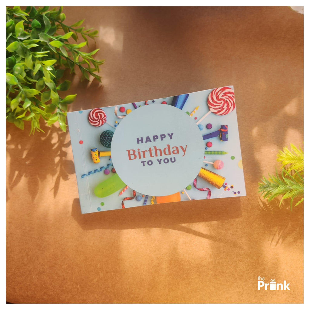 Birthday photo album on brown surface with images of colorful candies, confetti, and a message "HAPPY Birthday TO YOU." Green leaves frame the scene, creating a festive vibe.